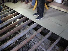 A photograph showing exposed wooden floor joists with a sheet of plywood being installed during a renovation.