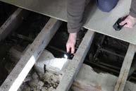 A close-up photograph showing a person using a flashlight to inspect wooden floor joists, likely during a structural survey or renovation.