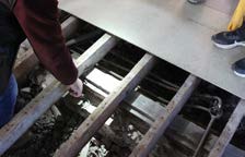 A close-up photograph showing exposed wooden floor joists and a sheet of plywood, with a person's arm and feet visible during construction work.