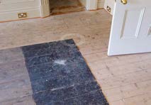 Interior view showing light wooden floorboards with a dark rectangular patch, white skirting boards, and a white door.
