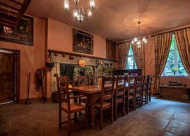Interior photograph of a traditional dining room featuring a long wooden table, stone flooring, and a tiled stove.