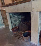 Interior photograph of a rustic room featuring a large stone fireplace with a wooden mantelpiece and traditional furnishings.