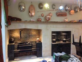 Interior photograph of a kitchen featuring a large brick fireplace with a range cooker and hanging copper cookware.