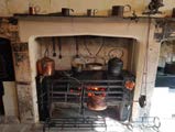 A photograph showing a traditional interior kitchen featuring a large stone fireplace surround with a black cast-iron range and hanging copper pots.