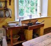Interior photograph of a room, likely a kitchen, featuring a wooden workbench and a window looking out onto greenery.