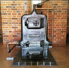 A photograph of a vintage Galloway's Patent Steam Engine boiler situated indoors against a brick wall.