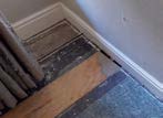 A close-up photograph showing an interior floor under renovation with exposed subflooring, wooden planks, and a skirting board.
