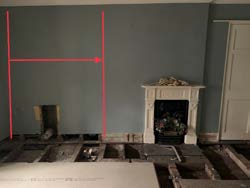 Interior photograph of a room undergoing renovation with a fireplace, exposed floor joists, and red measurement lines on the wall.