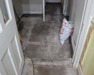 An interior photograph showing a room with unfinished concrete flooring and a bag of building materials, likely during renovation work.