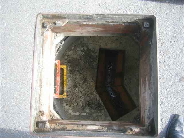 A close-up photograph of an open square manhole or inspection chamber with a yellow measuring tape visible inside.