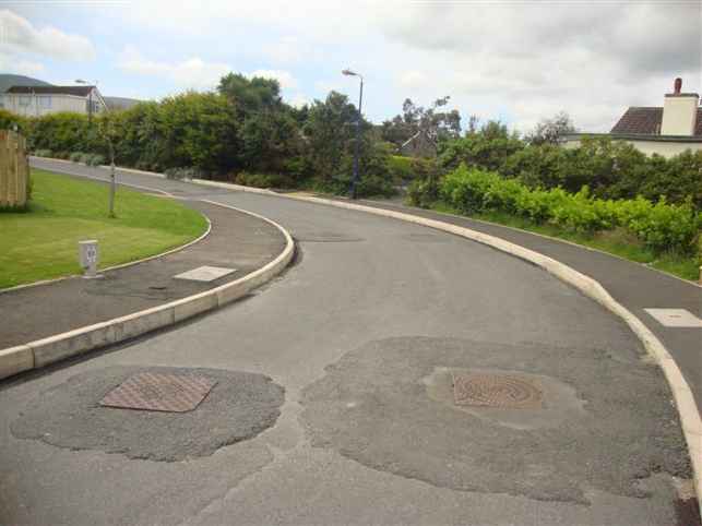 A photograph showing a curved paved road with concrete curbs, drainage access covers, and surrounding green landscaping.
