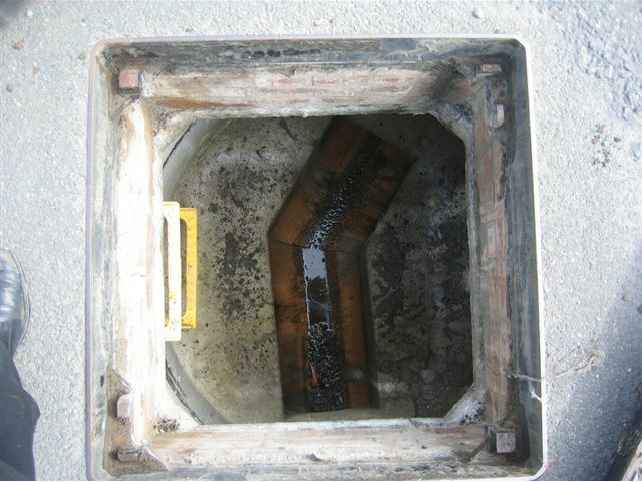 A photograph looking down into an open square manhole or inspection chamber revealing internal drainage pipes and a yellow ladder.