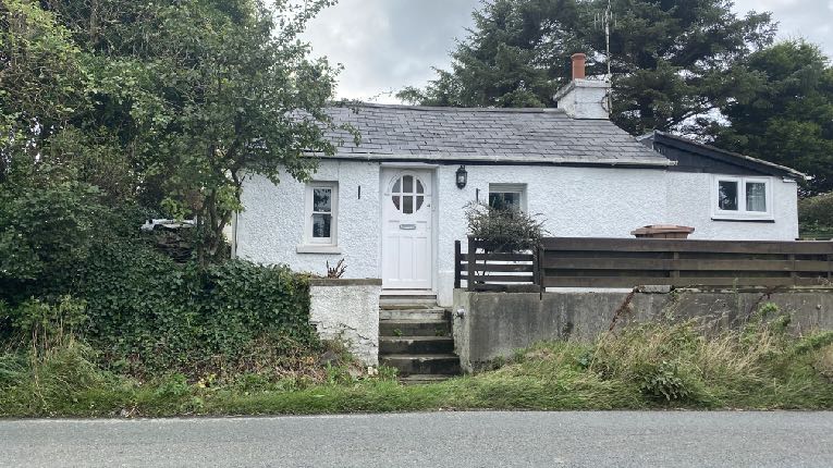 A photograph of a white single-story bungalow with a slate roof and a side extension, set amongst trees and greenery.