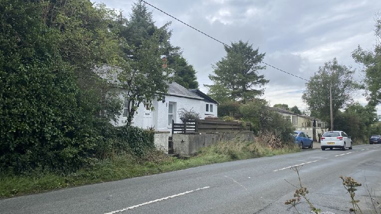 A street-level photograph showing a white single-story house (bungalow) situated beside a road, surrounded by trees and vegetation.