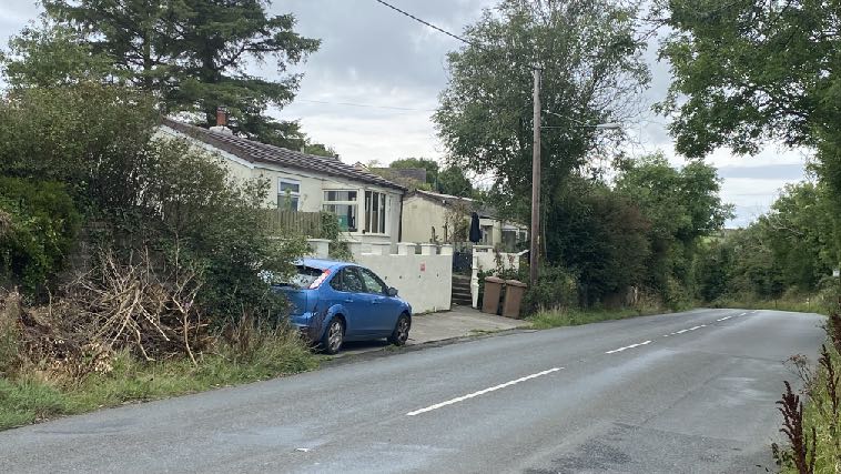 A street-level photograph showing a white single-story bungalow with a blue car parked in front, surrounded by trees and vegetation along a road.