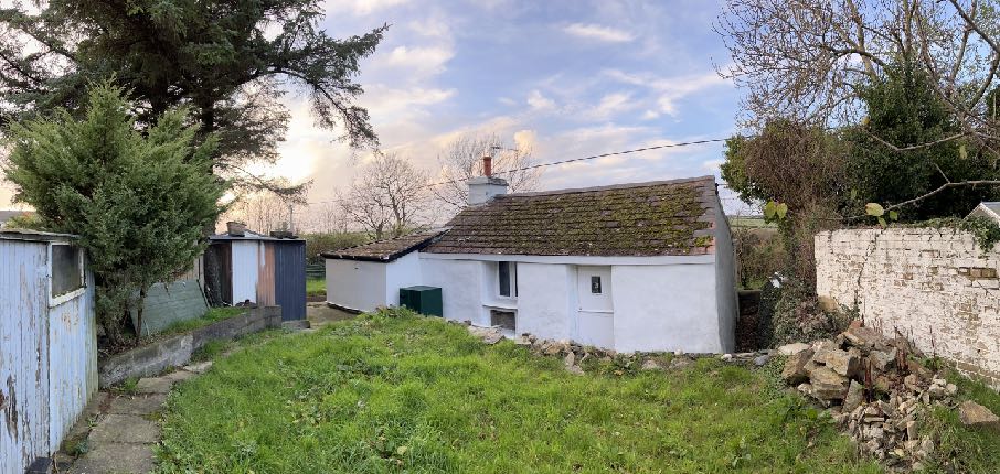 A photograph showing a white single-story cottage or bungalow with a tiled roof, situated in a rural setting with adjacent sheds and a stone wall.