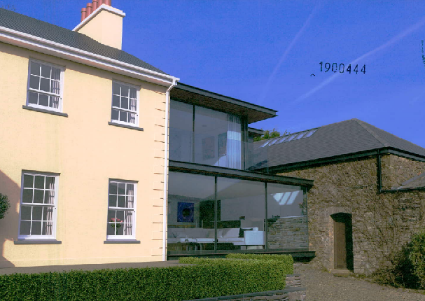 A photograph showing a cream-colored two-story house with a modern glass extension connecting it to an adjacent stone building under a blue sky.