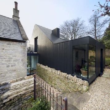 A photograph showing a modern dark-clad extension attached to a traditional stone building, featuring large glass windows and a stone wall boundary.