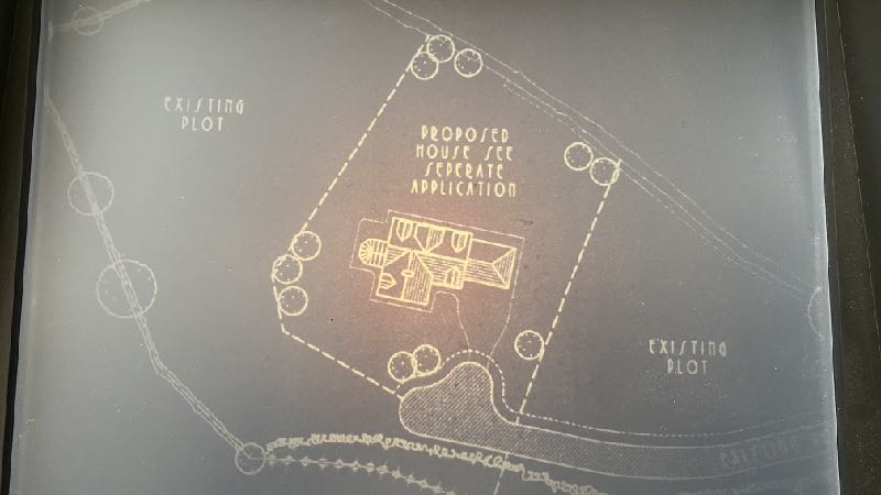 A site plan showing a proposed house footprint within a dashed boundary line, surrounded by trees and adjacent to existing plots.