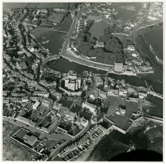A black and white aerial photograph showing a coastal town with a prominent castle structure and surrounding buildings.