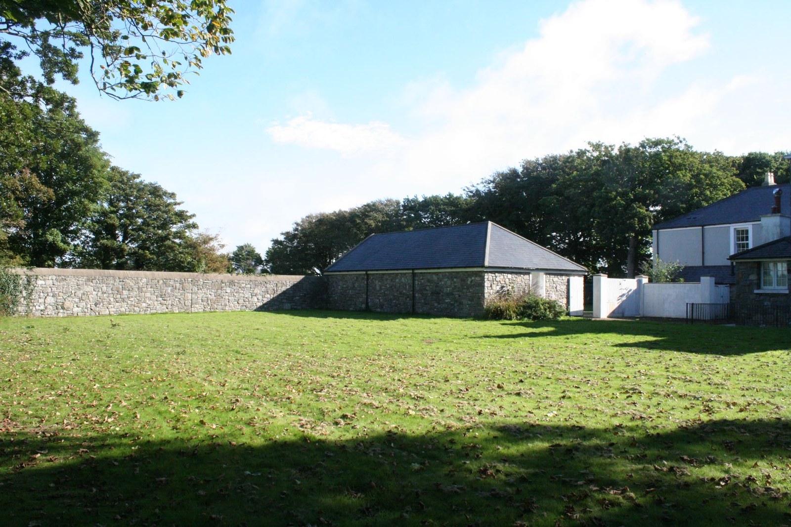 A photograph showing a grassy garden area with a stone outbuilding and a stone wall, with a white house visible on the right side.