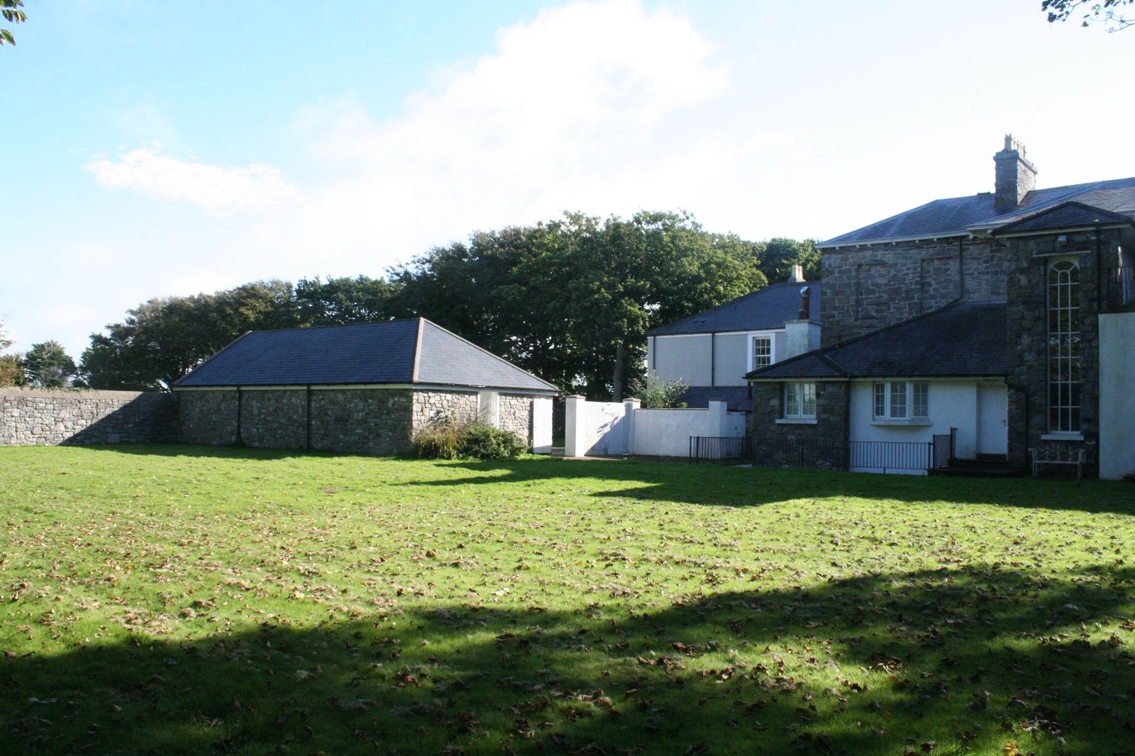 A photograph showing a large grassy garden area with stone buildings in the background, including a single-story outbuilding and a larger house.