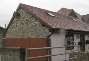 A photograph showing the exterior of a stone and rendered building with a tiled roof and skylights, viewed from behind a metal gate.