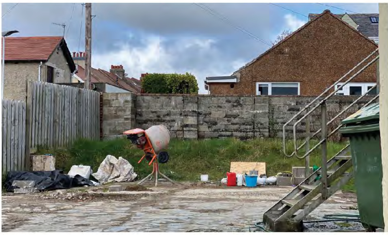 A photograph of a construction site or yard featuring a cement mixer, building materials, and a concrete block wall with residential houses in the background.