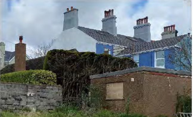 A street-level photograph showing a row of residential houses with pitched roofs and chimneys, partially obscured by hedges and a low wall in the foreground.