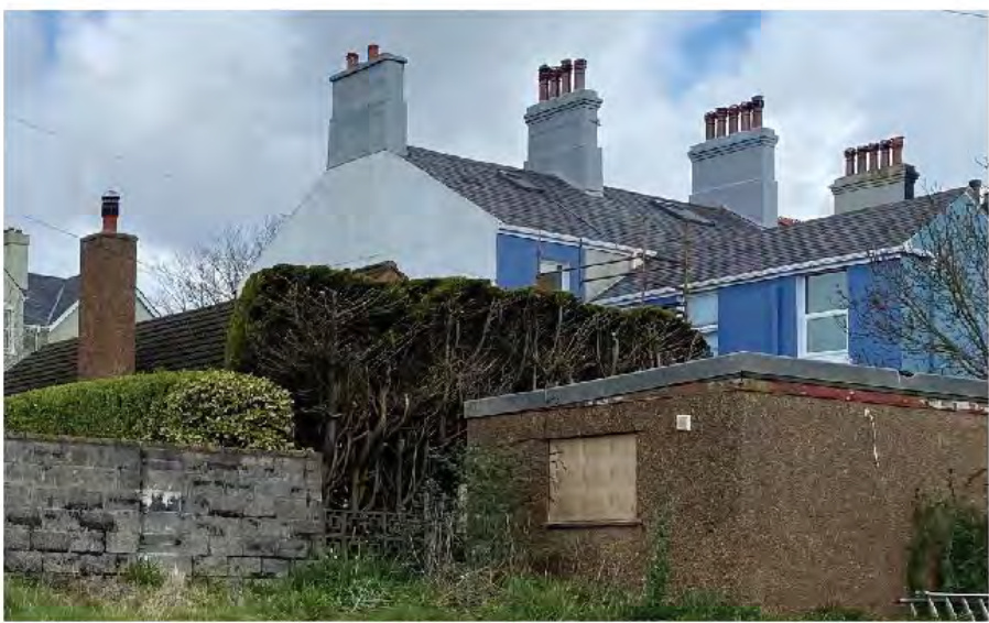 A photograph showing a row of terraced houses with blue and white facades behind a low brick wall and overgrown vegetation, with a small brick outbuilding in the foreground.