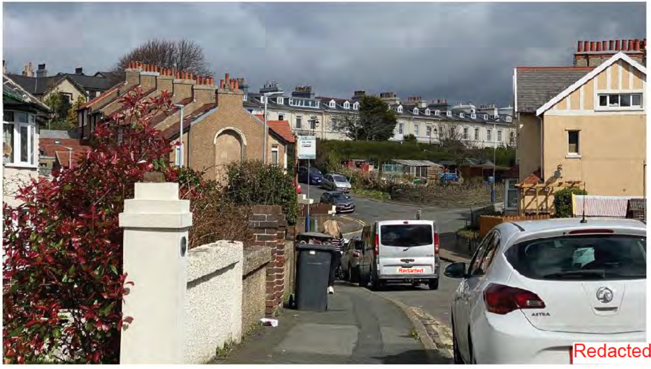 A street-level photograph showing a residential road with parked cars, brick walls, and terraced houses in the background under a cloudy sky.