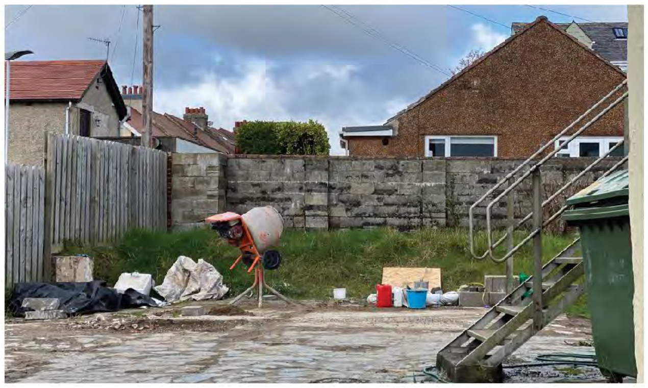 A photograph of a residential rear yard or construction site featuring a cement mixer, building materials, and boundary walls.