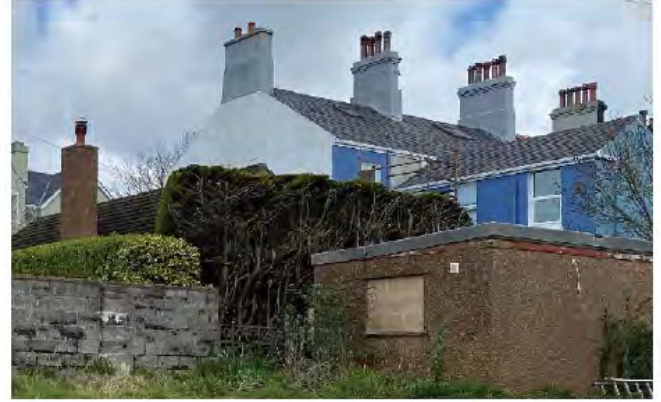 A photograph showing the rear or side elevation of a row of terraced houses with slate roofs and chimneys, viewed from behind a stone wall and hedge.
