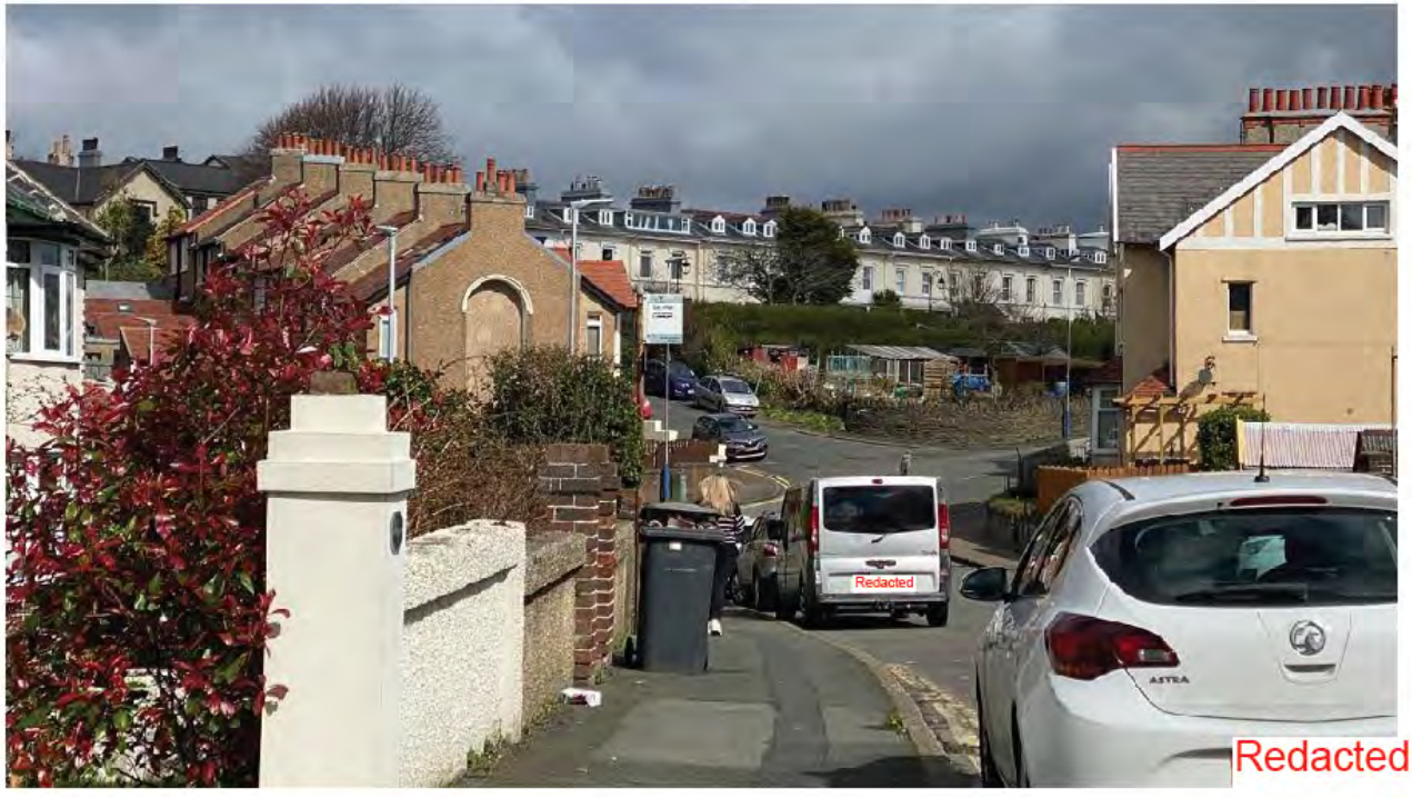 A street-level photograph showing a residential road with parked vehicles, a foreground wall, and a row of terraced houses on a hill in the background.