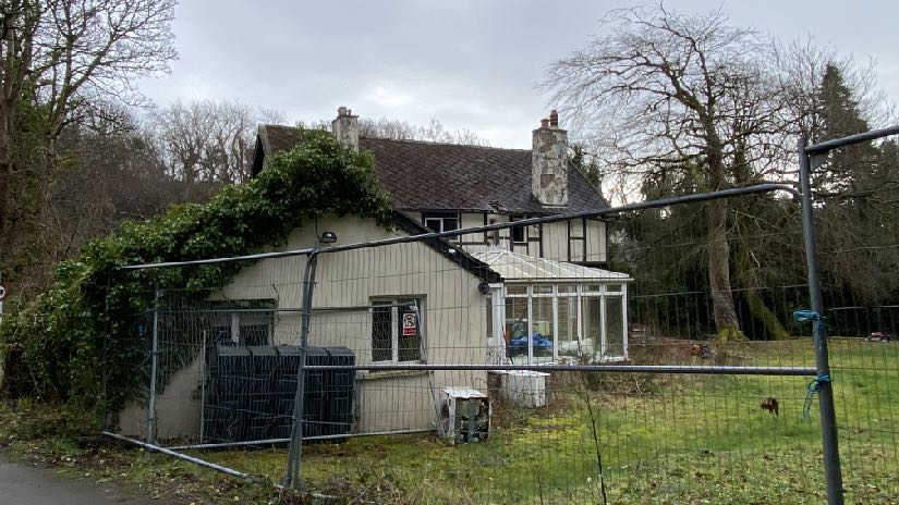 A photograph showing a detached house with a conservatory, viewed through a temporary metal fence in a green, rural setting.