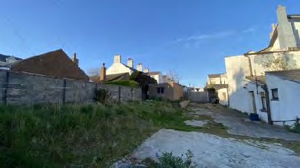 This photograph shows the existing site conditions, featuring an overgrown grassy area in the foreground, a stone boundary wall on the left, and existing white buildings with chimneys in the background.