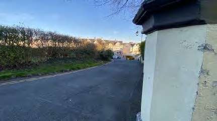A street-level photograph showing a paved access road leading towards a row of residential buildings, with a white boundary wall in the foreground.