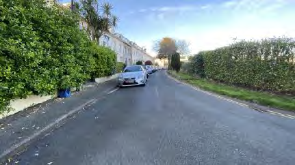 A street-level photograph showing a residential road with parked cars, green hedges, and a row of houses in the background.