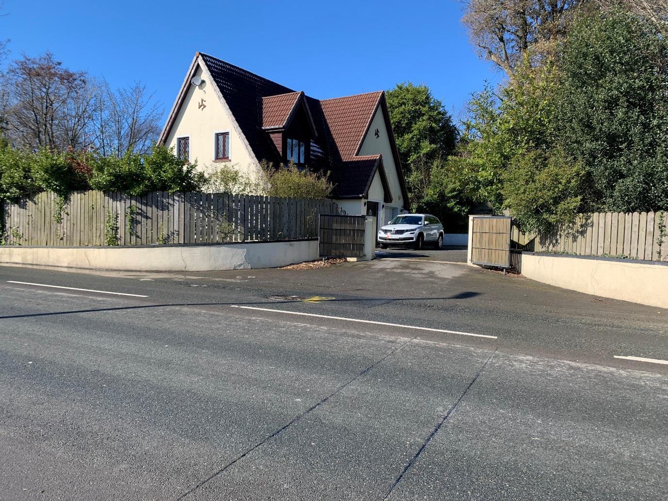 A street-level photograph showing a detached house with a steep gable roof, wooden boundary fencing, and a driveway with a parked car.