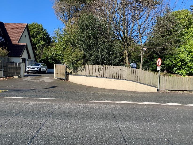 A street-level photograph showing a residential property entrance with a driveway, wooden fence, and retaining wall adjacent to a road.