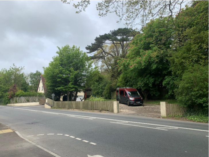 A street-level photograph showing a residential property with a driveway, wooden fencing, and surrounding trees.