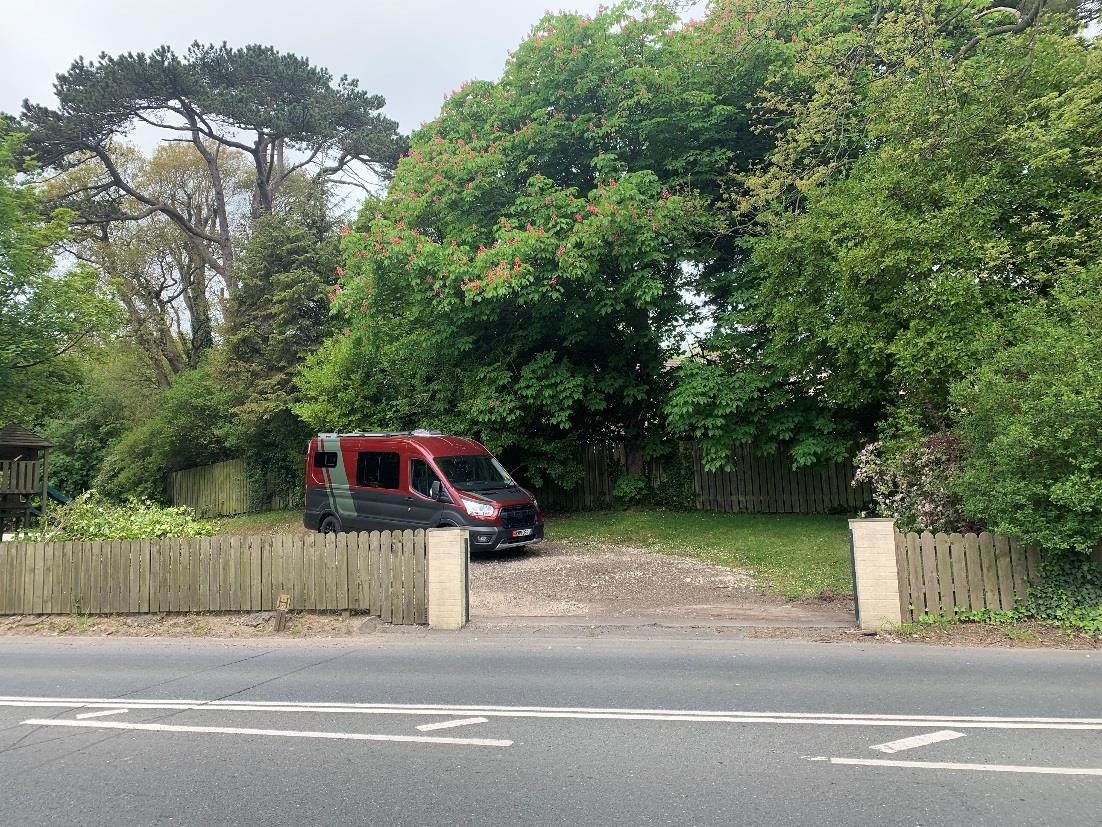A street-level photograph showing an existing gravel driveway entrance with a wooden gate and fence, where a red camper van is parked.