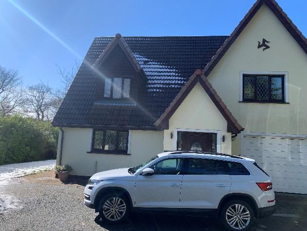 A photograph showing the front exterior of a detached two-storey house with cream walls and a dark tiled roof, with a white car parked on the gravel driveway.