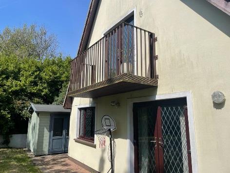 Exterior photograph of a two-story house featuring a wooden balcony, security grilles, and a small garden outbuilding.