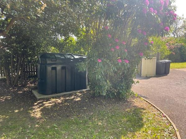 A photograph showing a large black plastic bin store situated on a concrete base next to a paved driveway and garden foliage.