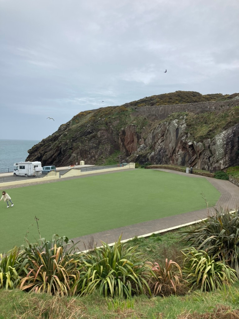 A photograph showing a large artificial green sports field situated next to the sea with a rocky cliff in the background and vehicles parked nearby.