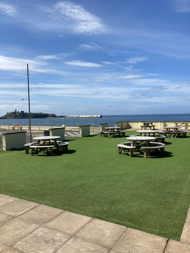 The image shows an outdoor patio area covered in artificial grass with wooden picnic tables, overlooking the sea and Peel Castle in the distance.