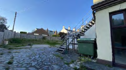 A photograph showing the rear exterior of a property featuring a cobblestone yard, an external metal staircase, and a green waste bin. A wooden fence and neighboring houses are visible in the background under a blue sky.