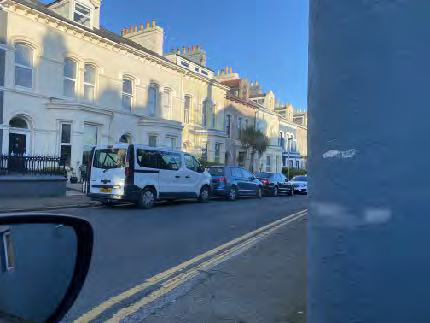 A street-level photograph taken from a vehicle showing a row of buildings and parked cars along a road with double yellow lines.
