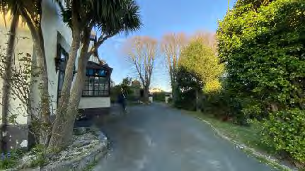 A street-level photograph showing a white residential building with a bay window on the left, surrounded by trees and a stone wall, with a road leading into the distance.